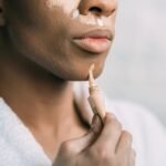 Crop anonymous African American man doing facial makeup with foundation cream as drag queen